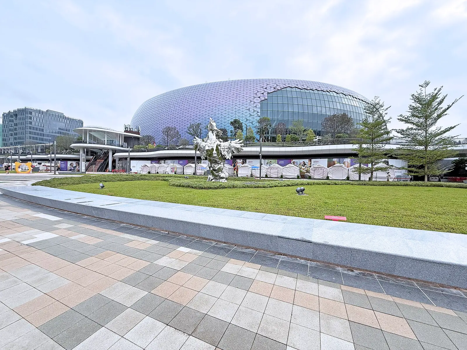 The domed Kai Tak Sports Park building seen across paved plaza and lawn